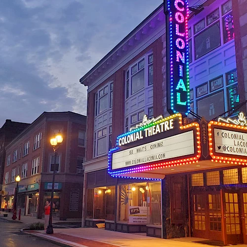 A neon-lit theater marquee on a street with brick buildings at dusk, showing &ldquo;Colonial Theatre&rdquo; signs and warm streetlights.