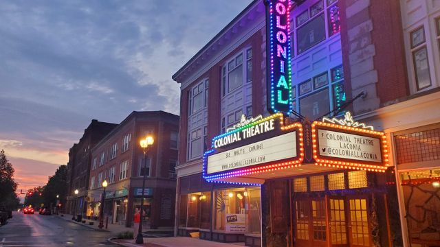 A neon-lit theater marquee on a street with brick buildings at dusk, showing &ldquo;Colonial Theatre&rdquo; signs and warm streetlights.