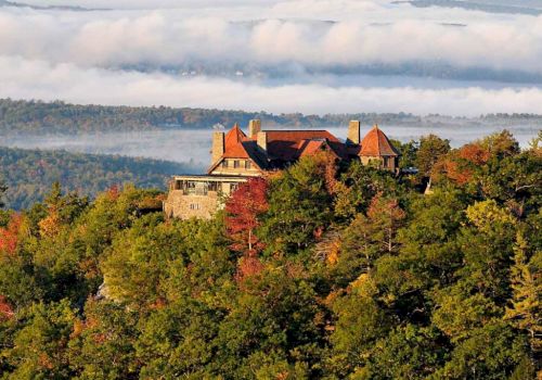 A castle sits atop a wooded hill, autumn leaves in orange and green, with rolling hills in the distance under a cloudy sky.