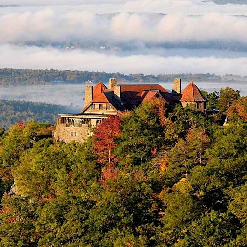 A castle sits atop a wooded hill, autumn leaves in orange and green, with rolling hills in the distance under a cloudy sky.