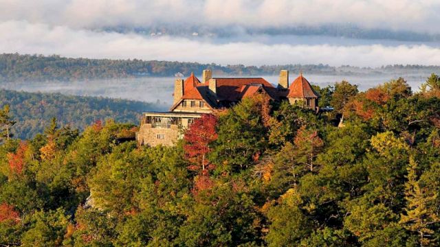 A castle sits atop a wooded hill, autumn leaves in orange and green, with rolling hills in the distance under a cloudy sky.