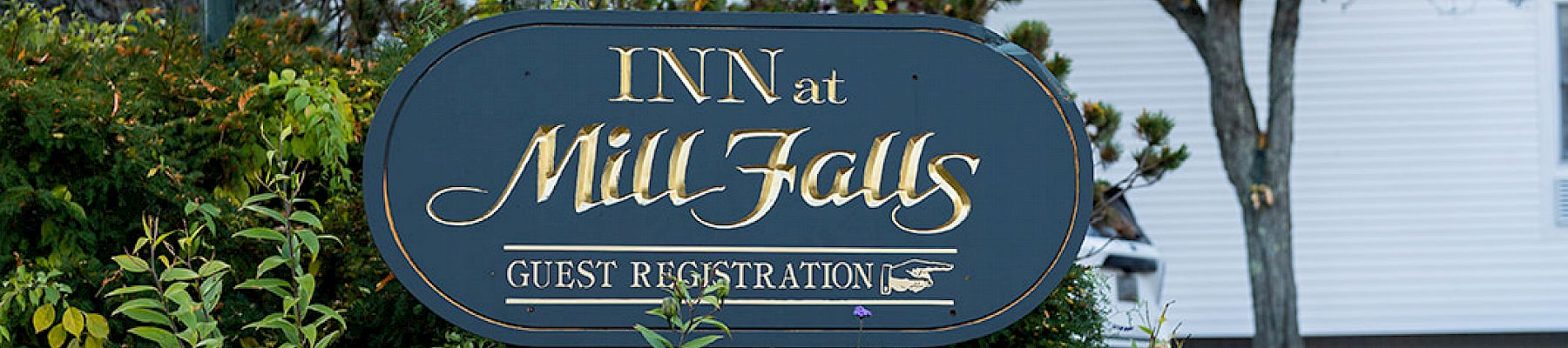 An inn sign with "Inn at Mill Falls" set amidst autumn foliage, featuring a landscaped garden with a white building in the background.