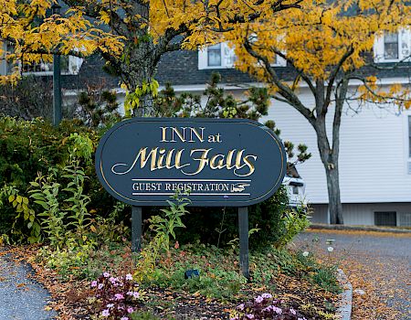 An inn sign with "Inn at Mill Falls" set amidst autumn foliage, featuring a landscaped garden with a white building in the background.