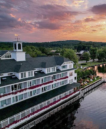 A lakeside hotel with a wraparound porch sits beside calm water at sunset, a pier extends into the lake, and tree-covered hills frame the scene.