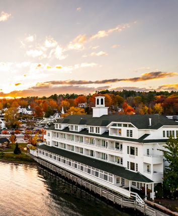 A riverside hotel and town with white buildings, autumn trees, and a glowing sunset sky.