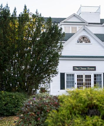 A white, two-story building with a gabled roof, trimmed windows, and a sign reading &ldquo;The &hellip; House&rdquo; behind a hedge and shrubs, in a garden setting.