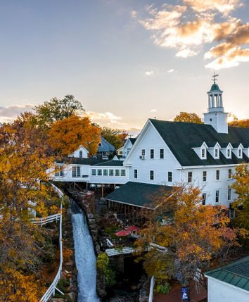 A white historic campus building with a tall bell tower, surrounded by autumn trees under a sunset sky.