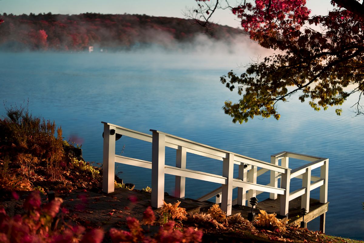 A lakeside view with misty blue water and a white railing deck, framed by autumn trees and a red-tinged sky.
