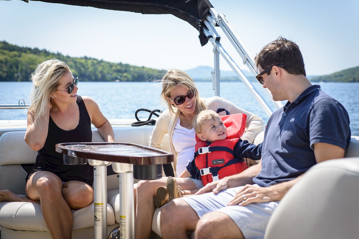 A family enjoys a sunny boat ride: two women, a man, and a child in a life jacket share laughs on a lake with trees in the background.