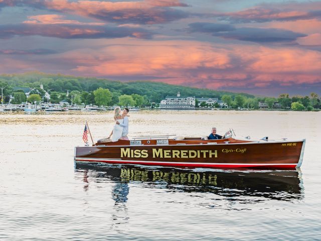 A couple stands on a wooden, vintage ChrisCraft boat named "Miss Meredith" on a calm lake, with a scenic shoreline and a colorful sunset sky in the background.