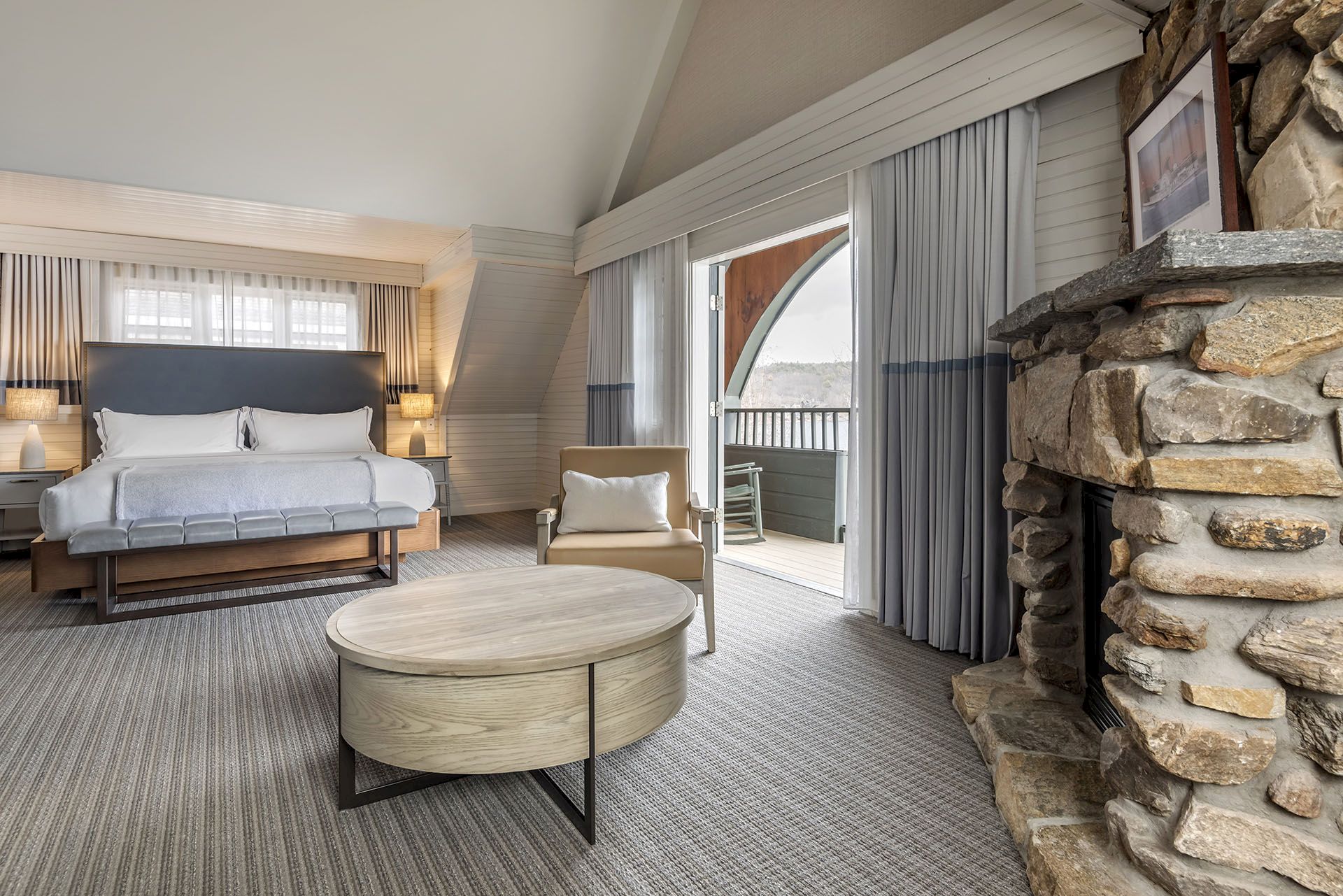 A spacious hotel suite with a white loveseat, a round coffee table, a stone fireplace, and a balcony view through sliding doors, warmly lit.