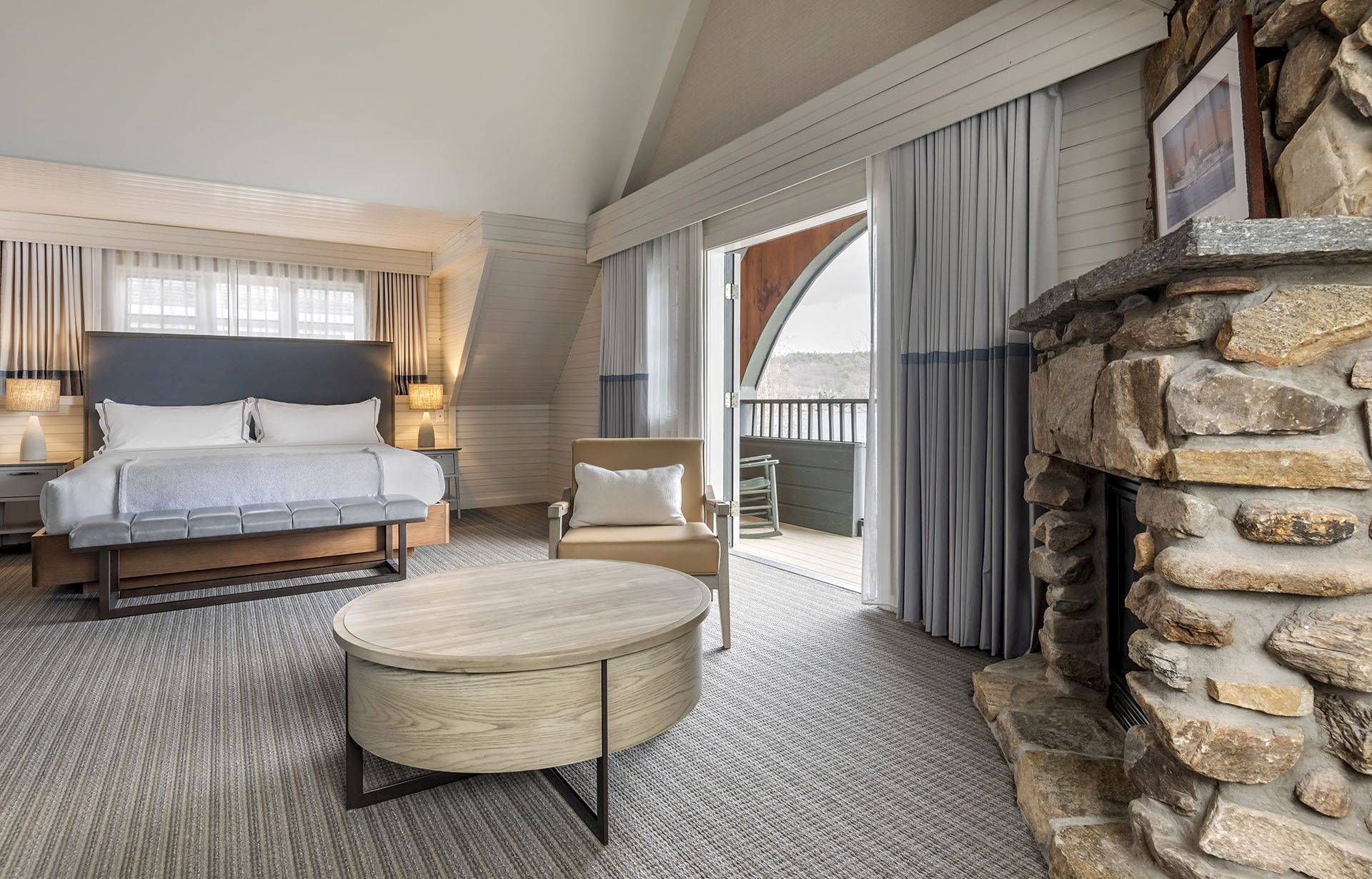 A spacious hotel suite with a white loveseat, a round coffee table, a stone fireplace, and a balcony view through sliding doors, warmly lit.