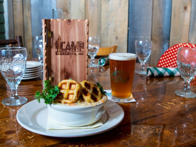 A table setting with a beer, a dish topped with mini waffles, fresh parsley, and a menu labeled "Camp." Glasses and napkins also visible.