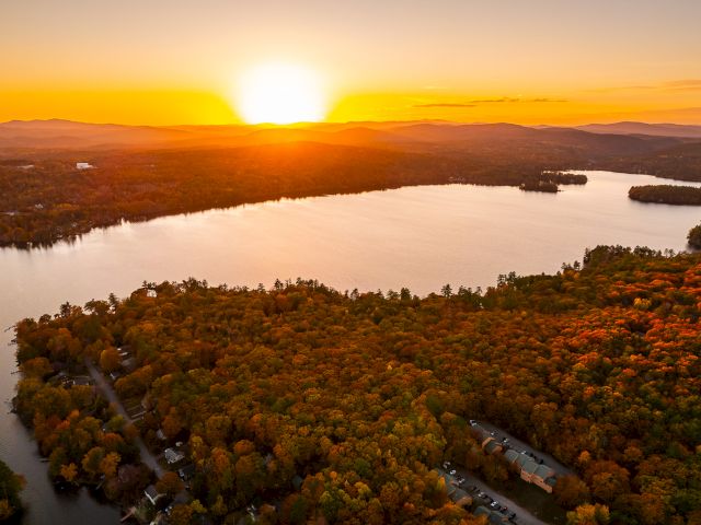 A golden sunset over a winding river and forested hills, with riverside homes and docks glowing in warm light.