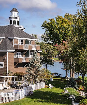 A lakeside house with a tower, green lawn, white benches, and trees by calm water under a blue sky.