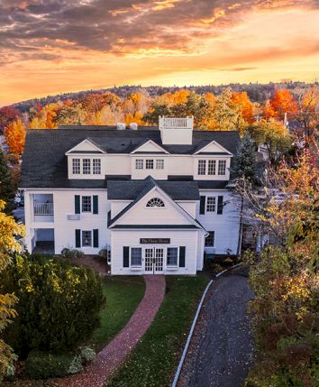 Aerial view of a large white mansion with a winding driveway, surrounded by golden autumn trees at sunset.