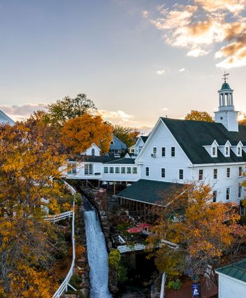 Autumn campus view with white colonial buildings and a church bell tower, plus a small waterfall under a sunset sky.