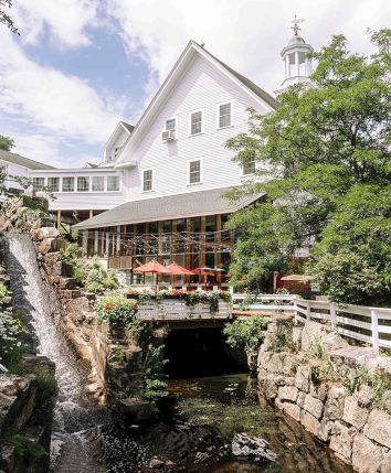 A picturesque white building over a rocky stream with a wooden deck, bridge railings, and leafy trees surrounding a sunny outdoor patio.