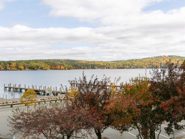 A scenic view of a calm lake with a wooden dock, surrounded by autumn-colored trees under a partly cloudy sky.