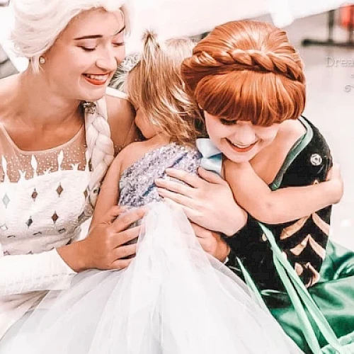 Two women in princess costumes hugging a child dressed as a princess, with joyful expressions.