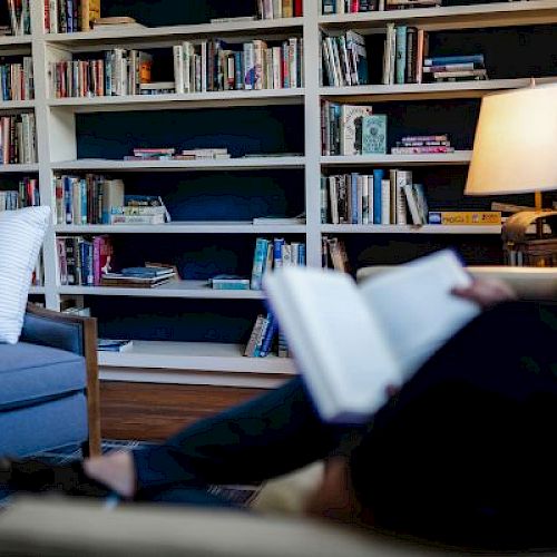 A person reads a book in a cozy room with full bookshelves, an armchair, and a lit lamp, creating a peaceful atmosphere.