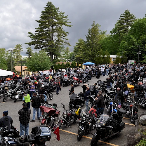 A large gathering of people and motorcycles in a parking area surrounded by trees under a cloudy sky.