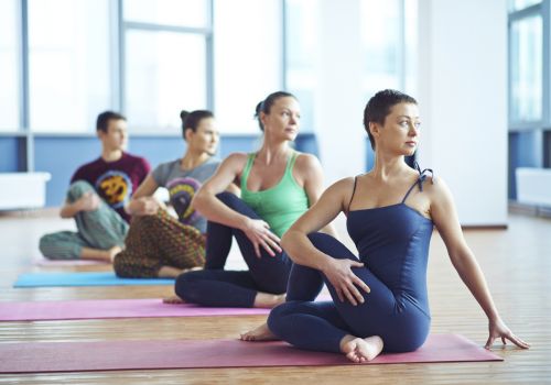 A group of people practicing yoga in a studio, sitting on colorful mats, engaged in a seated twist pose, with large windows in the background.