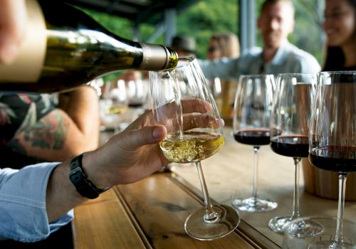 Friends toasting with wine glasses on a wooden table during a social gathering, pouring white wine while others chat in the background.