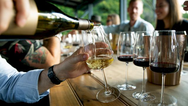 Friends toasting with wine glasses on a wooden table during a social gathering, pouring white wine while others chat in the background.