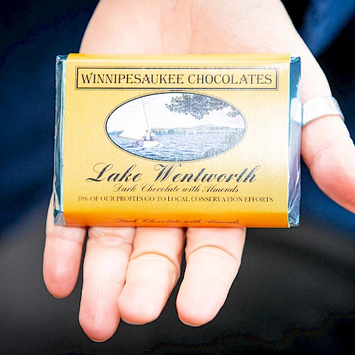 A hand holds a yellow bar of "Winnipeasaukee Chocolates," specifically "Lake Wentworth" with a sailboat illustration on the wrapper.
