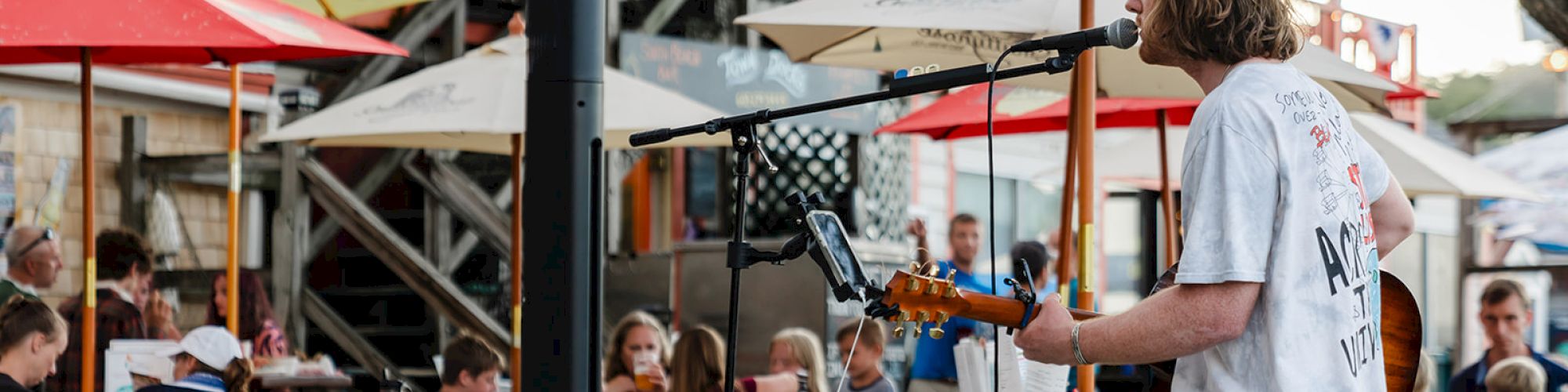 A musician performs with a guitar at a café with red umbrellas, as people dine and watch the entertainment.