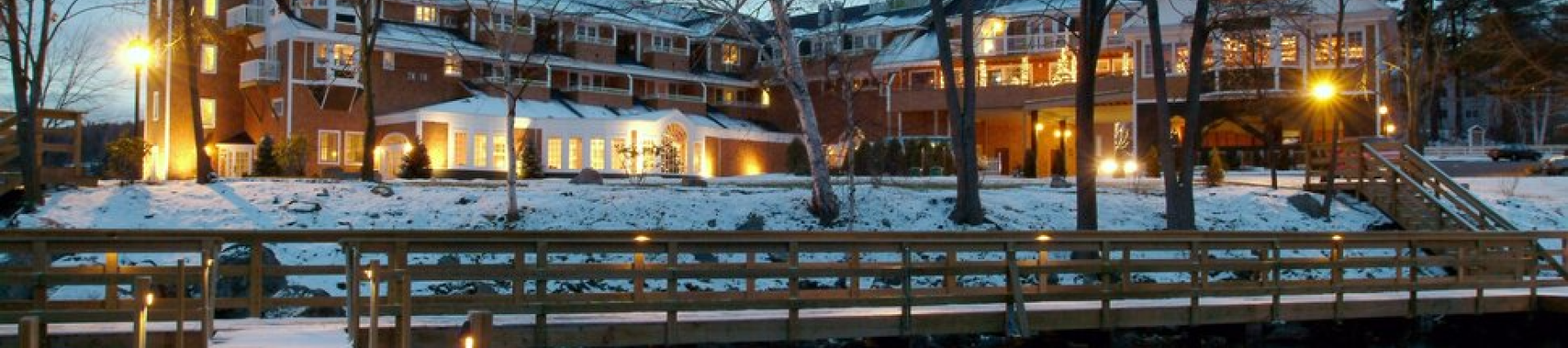 A building with a lit tower and glowing windows, surrounded by snow and trees, is viewed from a wooden dock over water at dusk.