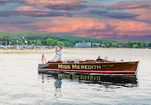 A couple stands on the "Miss Meredith" boat on a lake during sunset, with a picturesque landscape in the background.
