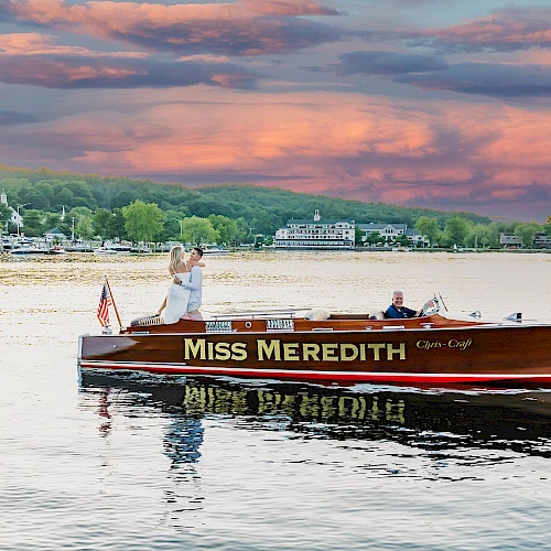 A couple stands on the "Miss Meredith" boat on a lake during sunset, with a picturesque landscape in the background.