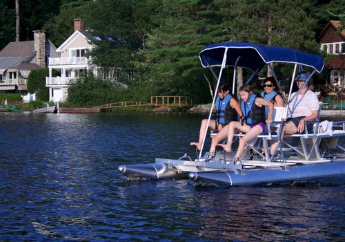 A family on a pedal boat enjoys a ride on a lake with waterfront houses and lush greenery in the background, all wearing life vests.