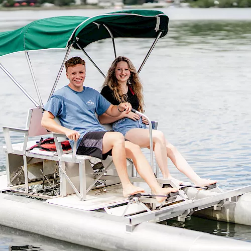 A couple smiles while riding a small boat on a lake, wearing life jackets and sheltered under a green canopy.