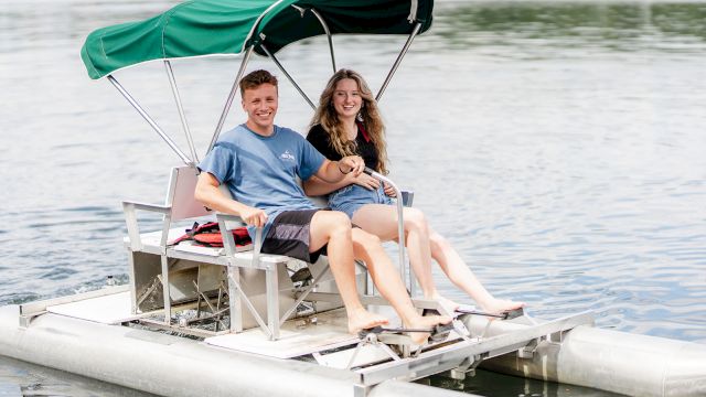 A couple smiles while riding a small boat on a lake, wearing life jackets and sheltered under a green canopy.