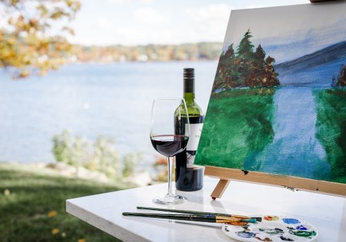 A bottle of red wine, a glass, and a painter&rsquo;s setup with an outdoors landscape painting while a lake and trees glow in the background.