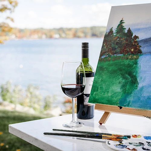 A bottle of red wine, a glass, and a painter&rsquo;s setup with an outdoors landscape painting while a lake and trees glow in the background.