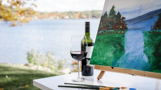 A bottle of red wine, a glass, and a painter’s setup with an outdoors landscape painting while a lake and trees glow in the background.