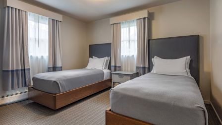 Two neatly made twin beds with gray sheets and white pillows sit in a bright hotel room, sharing a nightstand between them.