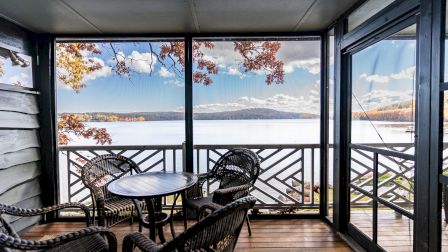 A screened balcony with wicker furniture overlooks a calm lake and distant mountains, framed by fall leaves and a wooden railing.