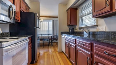 A modern kitchen with red wooden cabinets, black granite countertops, stainless steel appliances, and a small dining area visible in the back.