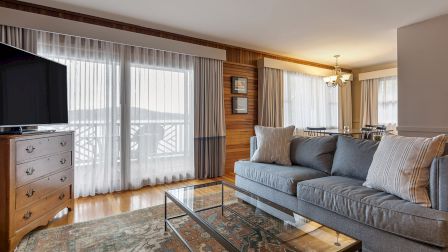 A bright, cozy living room with a gray sofa, glass coffee table, wooden floors, large windows with sheer curtains, and a flat-screen TV, ending here.