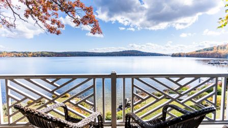 A peaceful lakeside view from a deck with wicker chairs, railing, calm water, blue sky, and autumn trees.