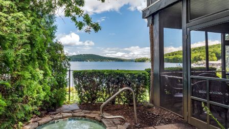 The image shows a small hot tub surrounded by greenery, overlooking a lake and hills, with a screened porch to the right.