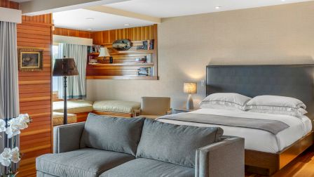 A modern bedroom with a couch, bed, wooden accents, a lamp, and a small desk area with shelves.