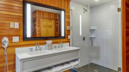 This image shows a modern bathroom with wooden walls, a vanity with mirror and lights, towels, and a glass-enclosed shower space.