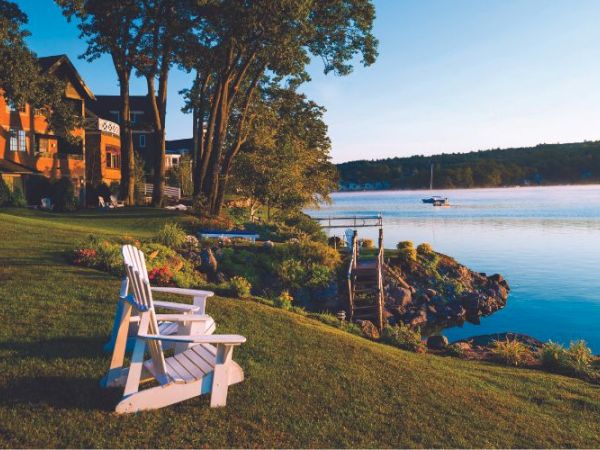 Lakeside scene with Adirondack chairs on a grassy lawn, houses to the left, dock by the water, and a sailboat in the calm lake.