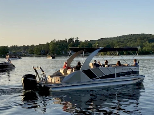 People are enjoying a ride on a pontoon boat on a lake, with trees and hills in the background during a sunny day.